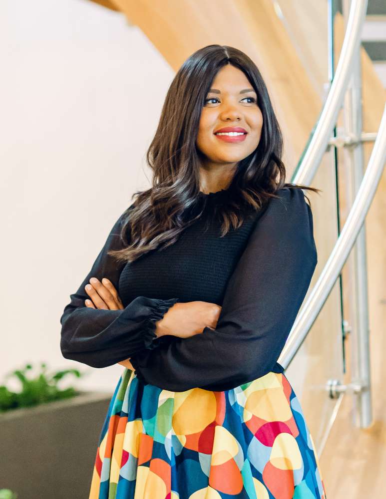 Mante Molepo is weating a black blouse and a multicoloured circular patterned skirt. She is smiling, with her arms folded, standing at the bottom of the stair-case with a white wall and small green plant behind her to the left.