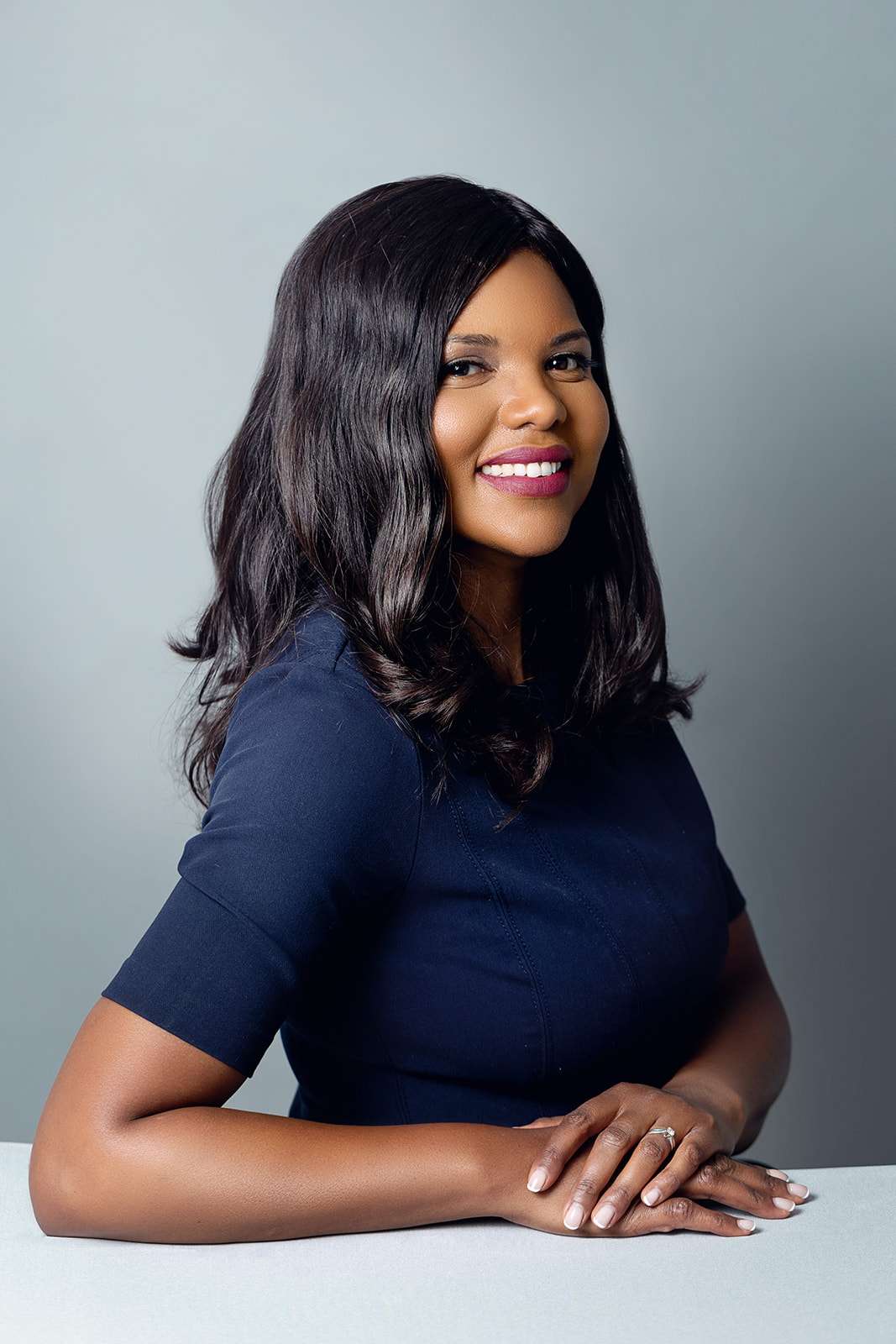 Mante Molepo standing and leaning on a desk, smiling and wearing a navy blue dress.