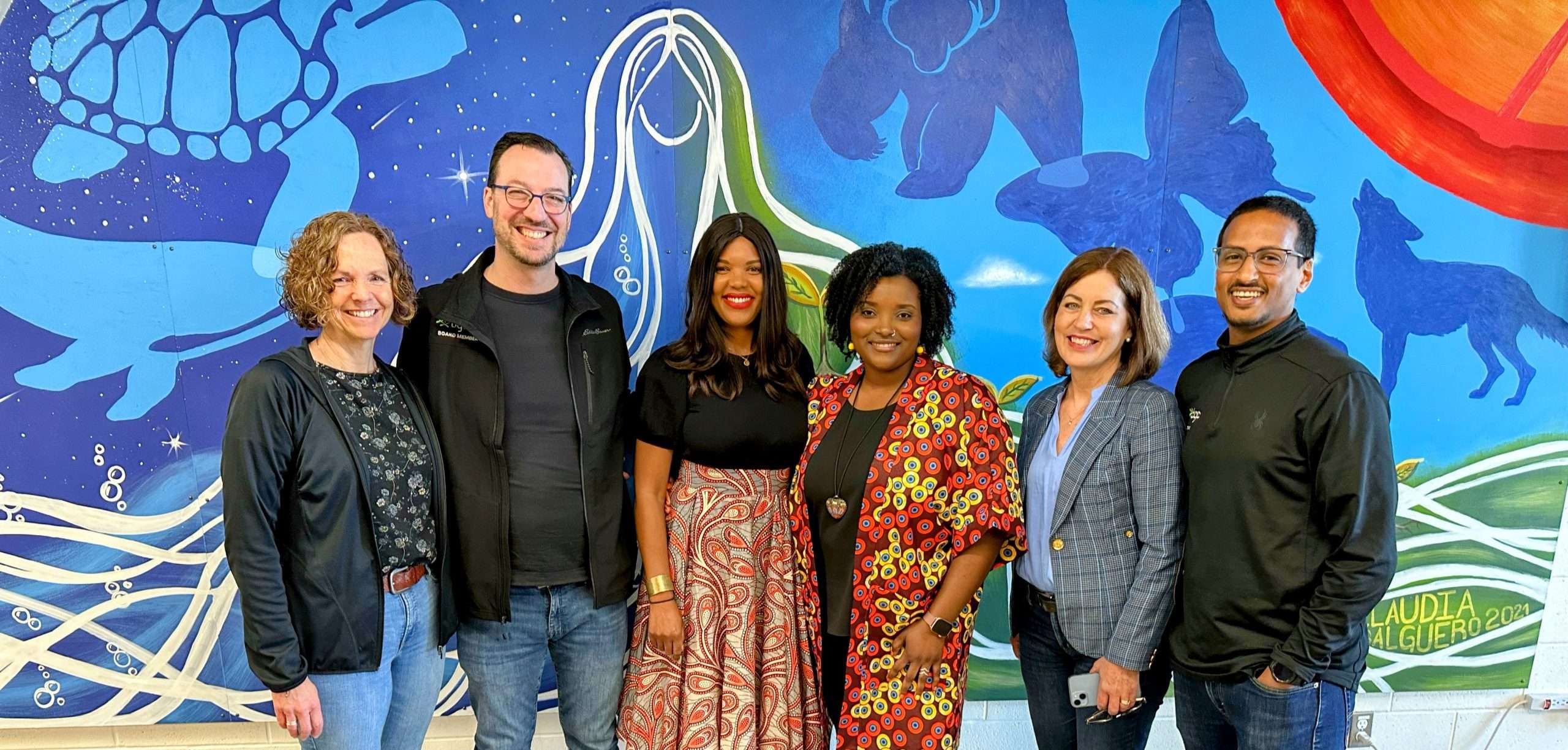 Mante stands in the middle of 5 other people smiling and wearing an orange patterned skirt and black blouse while standing in front of a brightly coloured sea life mural.