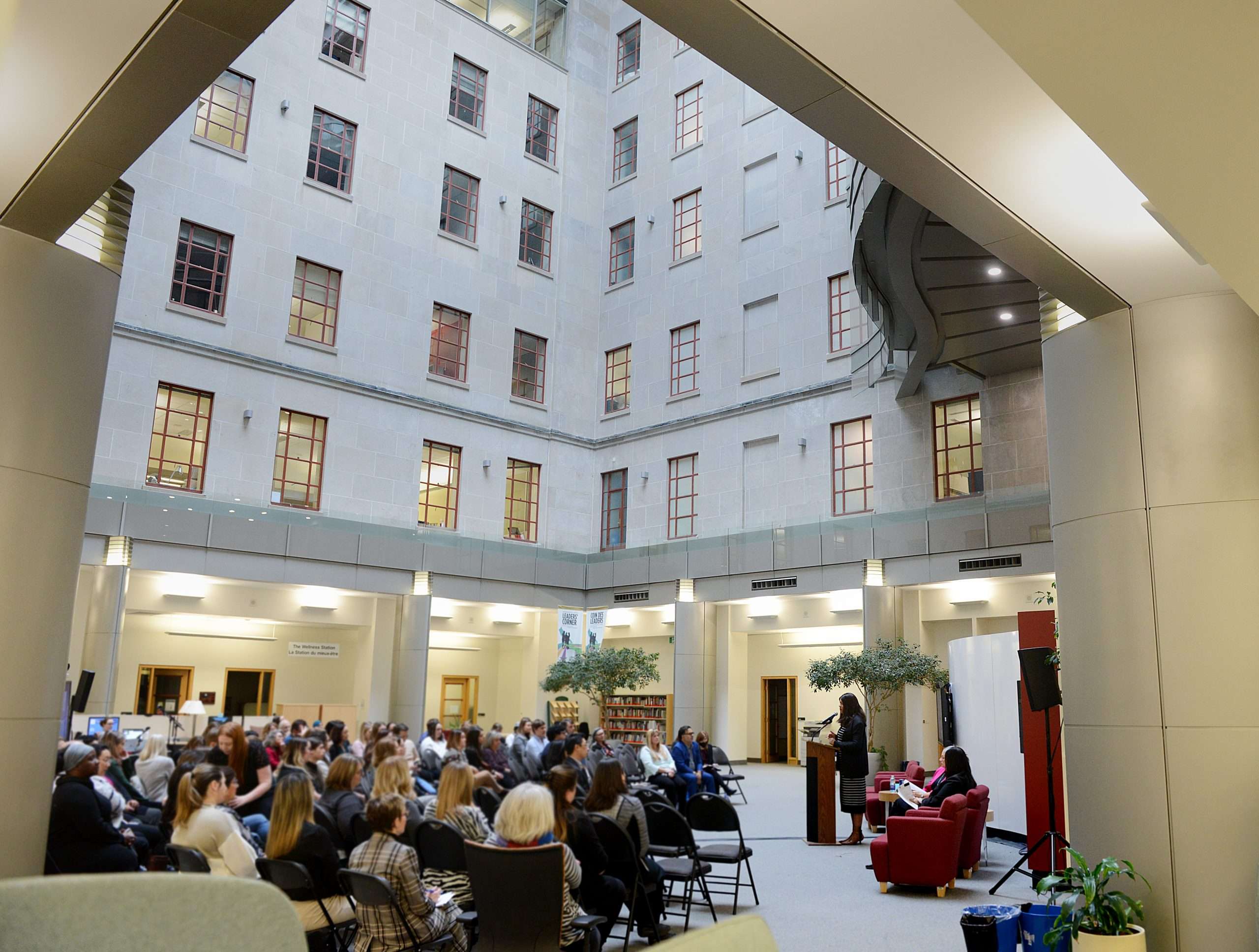 Mante presents in front of a podium, next to an artificial tree and in front of red couches. She addresses a group of people in an open concept space. The space has grey walls and brown paned windows overhead.
