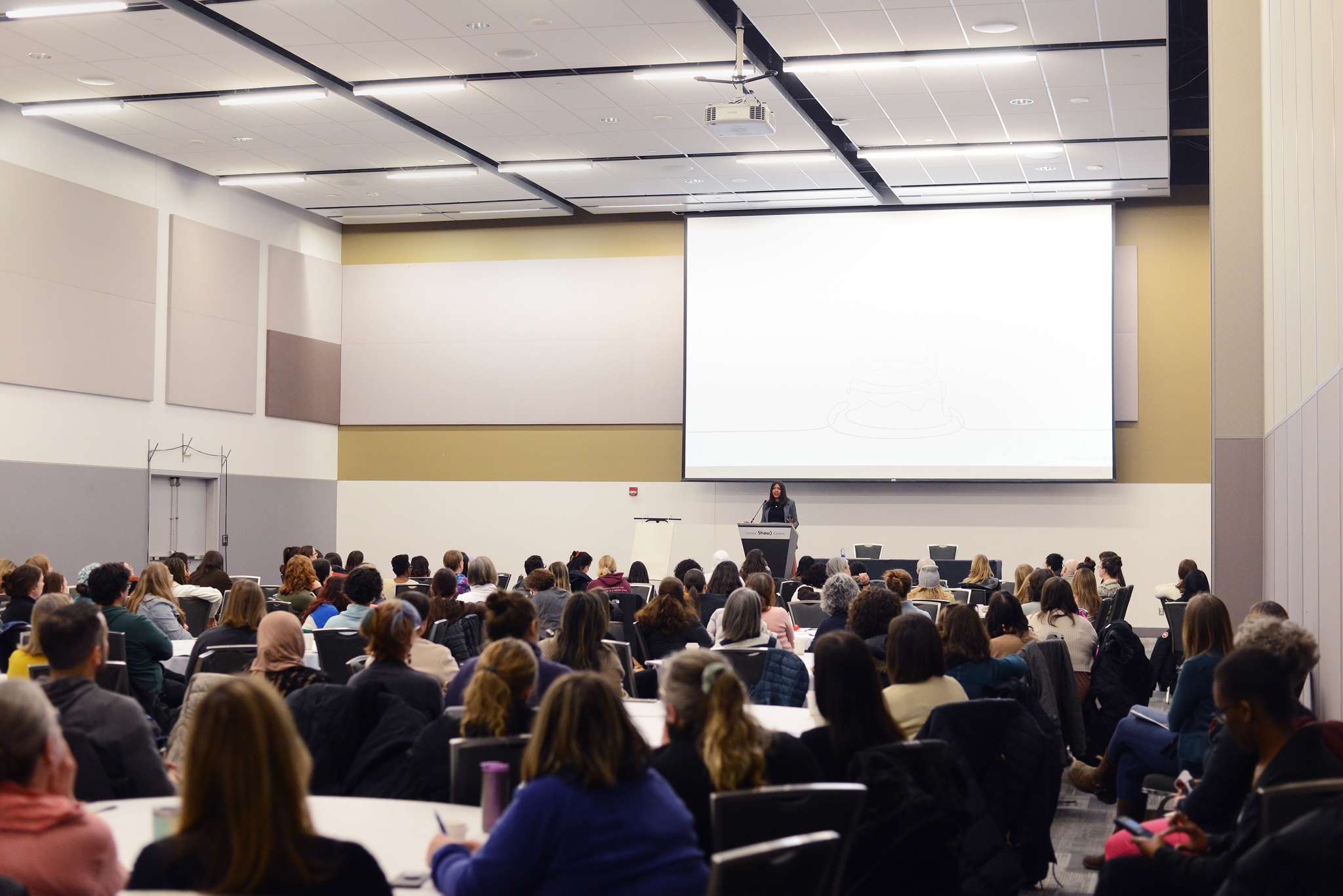 Mante is standing in front of a podium in the middle of a large room. Behind her is a white and blank presentation screen. In front of her is a room full of participants seated around tables. Some people are seated along the walls.