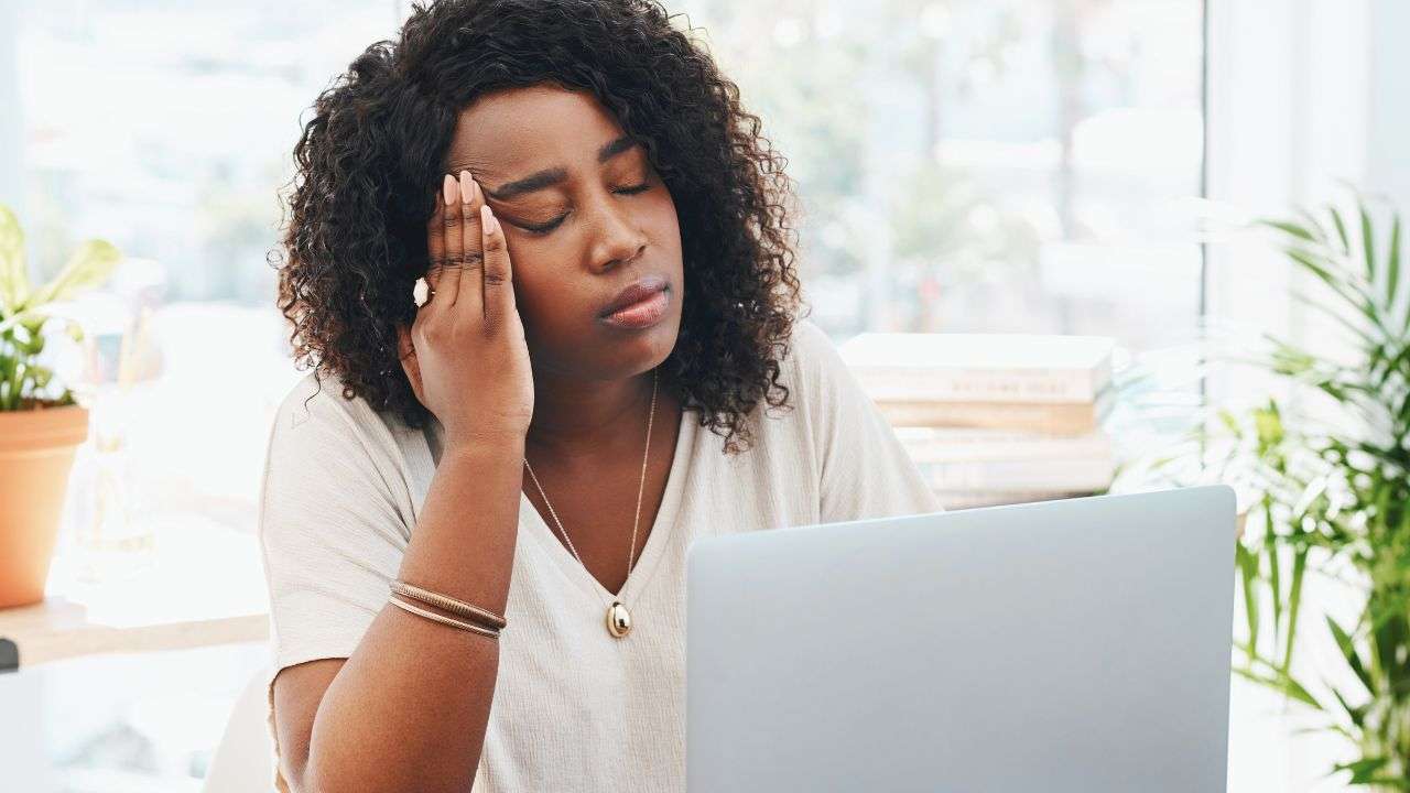 A Black woman sits at her laptop, holding her head as if to be stressed.