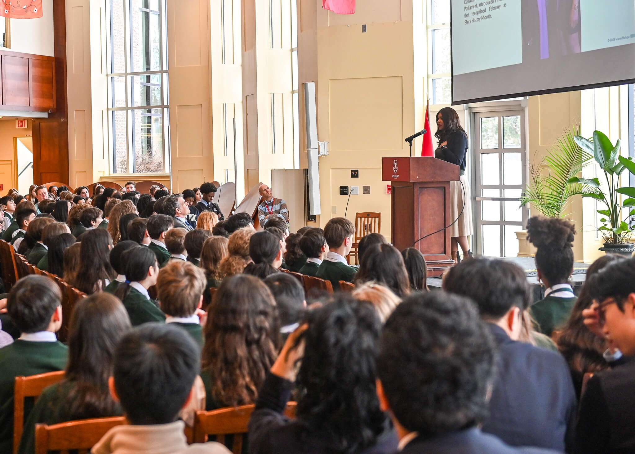 Mante Molepo keynote at Ashbury College on Ubuntu, Black History Month, and building inclusive student communities. Mante stands at a podium, speaking to an audience of students and faculty. Behind her are large glass doors, and a presentation screen hangs from the ceiling.
