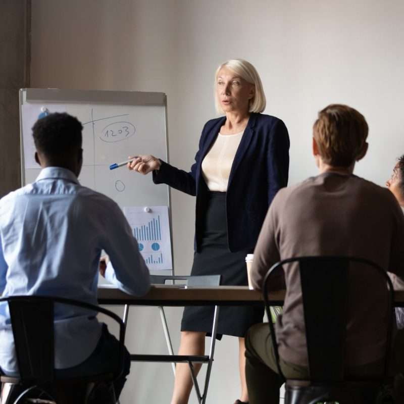 A woman is standing around a meeting table speaking to her leadership team and holding a pen while pointing to a flip chart.